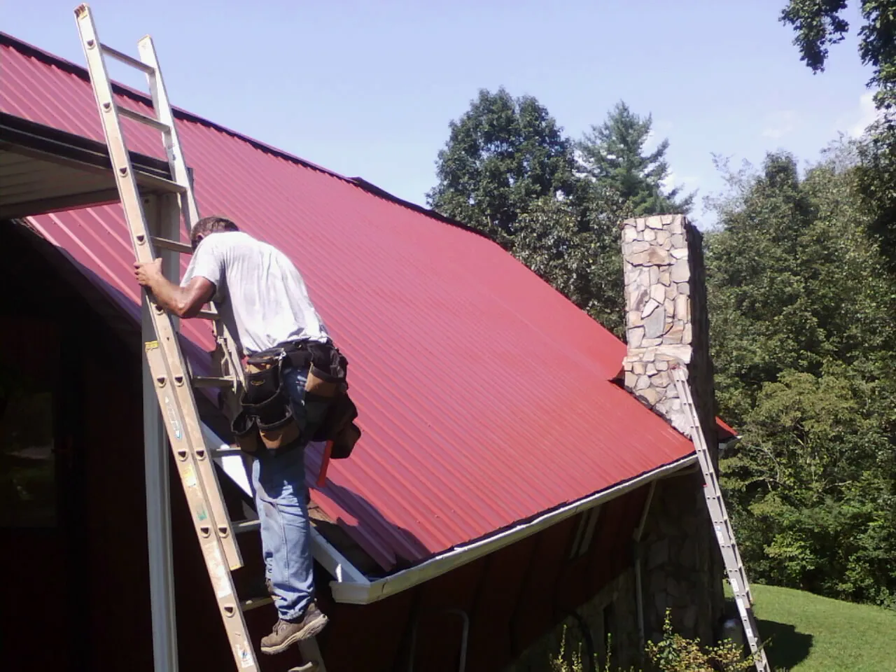 Roofer on a ladder beside a red metal roof and stone chimney — Soffit & Fascia Repair by Roofing Brothers in Little River
